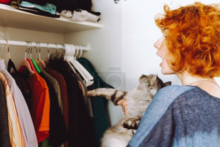 Red-haired curly teenage girl holds large gray Maine Coon cat in arms, choosing things from closet. spending time with pet. cat sits funny on girls shoulders
