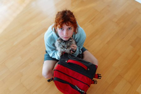 portrait red-haired teenage girl sitting on parquet floor, with suitcase, along with gray fluffy cat, in an empty apartment. concept moving, rent, student life