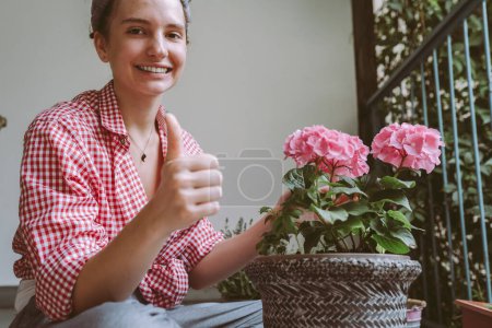 mujer joven, adolescente, atractiva, en camisa a cuadros, se sienta en el balcón, terraza, cuida la planta en maceta, trasplanta hermosa hortensia con pétalos rosados
