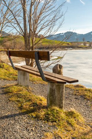 Photo for Einsiedeln area, Canton Schwyz, Switzerland, February 20, 2023 Empty bench at the coast of the frozen lake Sihlsee on a sunny day - Royalty Free Image