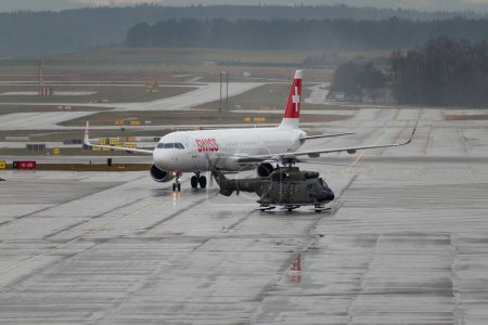 Photo for Zurich, Switzerland, January 17, 2024 Swiss Air Force Super Puma helicopter and an Airbus A321-212 jet on the apron in the rain during the world economic forum in Davos - Royalty Free Image