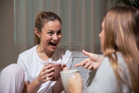 Photo for Happy excited woman talking with friend on sofa at night. Surprised sister receiving good news while having cup of coffee at home. - Royalty Free Image