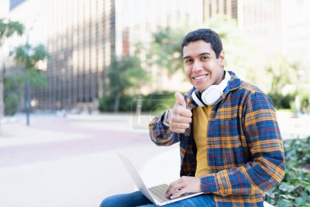 Photo for Portrait with copy space of a successful latin college student using laptop gesturing with thumb up - Royalty Free Image