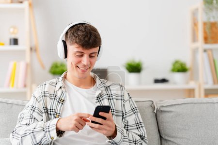 Teenager boy using music application on phone in the living room. Young man choosing song.