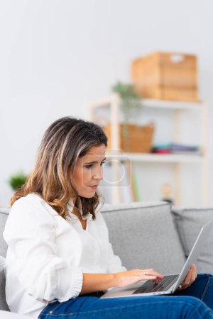 Vertical photo of a mature causal woman using laptop working from home on the sofa