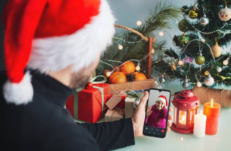 Téléchargez les photos : Appel vidéo à des amis la veille de Noël. Un homme appelle parents et amis sur un smartphone. La fille a répondu à l'appel vidéo et agite la main. En arrière-plan se trouve un arbre de Noël et la décoration. - en image libre de droit