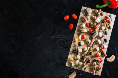 Téléchargez les photos : Snack skewers on a wooden tray on a black concrete table. Appetizers with mozzarella, ham, olives, cucumbers, tomatoes. - en image libre de droit