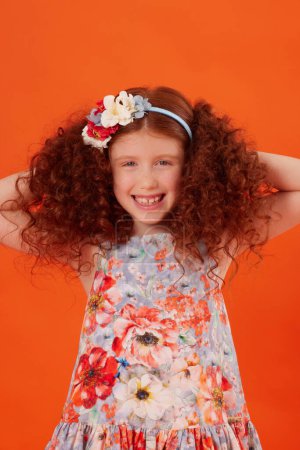 Smiling Redhead Girl with Freckles in Blue Floral Dress on Orange Background