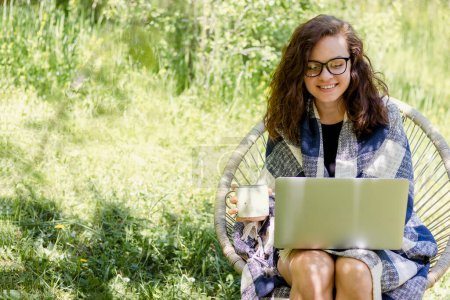 Photo for Happy young adult curly-haired woman wearing glasses uses a laptop while sitting in a green garden in the countryside. - Royalty Free Image