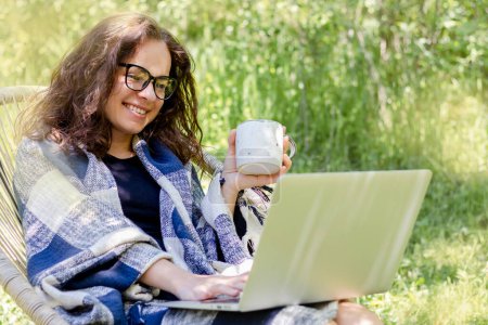 Foto de Woman wrapped in plaid using her laptop and drinking coffee in the garden. - Imagen libre de derechos