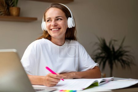 Photo for Happy female student in headphones sitting at desk at home doing homework in workbook. - Royalty Free Image