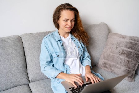Téléchargez les photos : Femme brune en chemise denim assise sur un canapé à la maison et utilisant un ordinateur portable. Achats en ligne. Travailler à domicile. - en image libre de droit