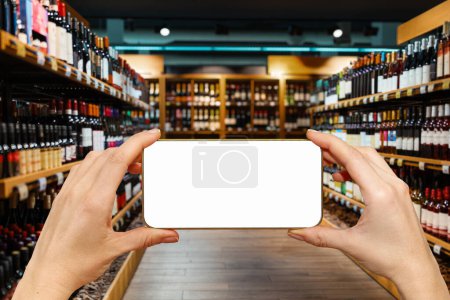 Foto de Teléfono inteligente irreconocible con pantalla en blanco en las manos delante de los estantes con botellas de vino en una licorería. - Imagen libre de derechos