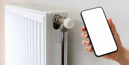 Remote control of domestic heating panel with smart phone and mobile app. Mobile phone with blank screen against white heating convector radiator.