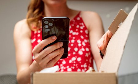 Femme en robe rouge d'été photographiant son achat en ligne dans un carton. Achats en ligne et partage de l'expérience consommateur. 