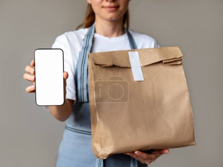 Bannière maquillable pour application mobile de commande et de livraison de nourriture. Serveuse tient le téléphone portable avec écran vide et sac en papier pour le repas à emporter dans ses mains.