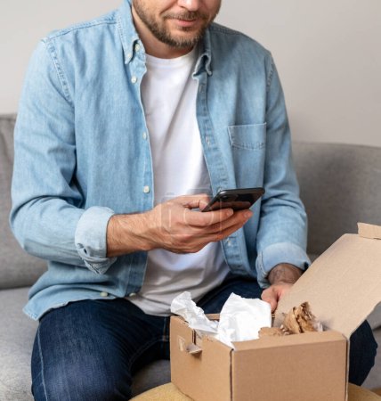 Homme dans une chemise en denim déballe un paquet et prend une photo de son achat, commande Internet. Personne partageant des commentaires avec la boutique en ligne à l'aide de son smartphone.