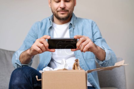 Un homme en jean déballonne un colis et prend une photo de son achat, commande internet. Personne partageant des commentaires avec la boutique en ligne à l'aide de son smartphone.