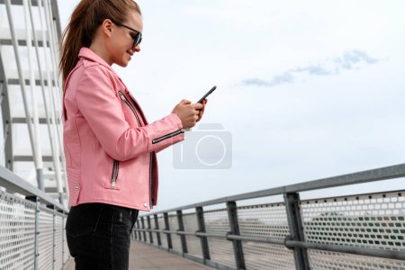 Photo for Stylish urban woman in pink jacket and sunglasses messaging online with her smartphone while walking outdoors in city. - Royalty Free Image