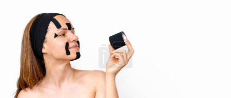 Woman beauty model with kinesio facial tapes on her face looks at roll of k-tape in her hand and smiling in front of white background with copy space.