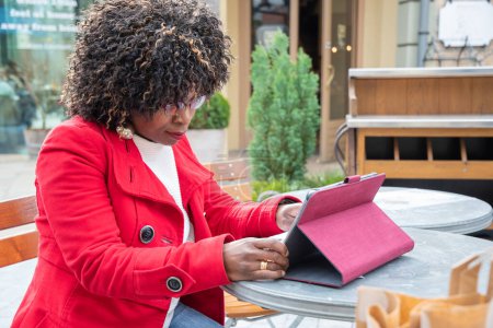 Foto de Una joven afroamericana con un abrigo rojo se sienta en una mesa en un café y la cuenta de comprobación después de comprar con una tableta, crisis de problemas financieros, la planificación adecuada de los gastos, foto de alta calidad - Imagen libre de derechos