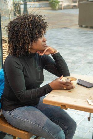 Foto de Pensive african american woman sitting in a cafe and looking out the window with a cup of hot, coffee or resting from household chores alone, High quality photo - Imagen libre de derechos