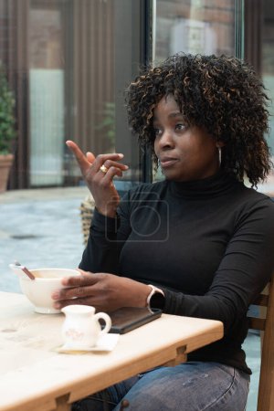 Foto de African american woman in red coat talking on smartphone near brick wall, technology use concept, wi-fi connection. High quality photo - Imagen libre de derechos