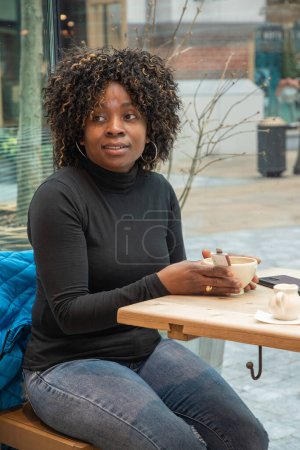 Foto de Mujer afroamericana sin maquillaje se sienta en un café por la mañana, después de haber despertado, y bebe café, Foto de alta calidad - Imagen libre de derechos