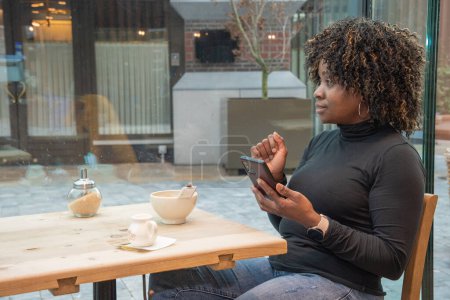 Foto de Joven afroamericana mujer sentada en un café con una taza de café caliente, viendo una fuente de medios sociales en un teléfono inteligente, utilizando la tecnología, foto de alta calidad, foto de alta calidad - Imagen libre de derechos