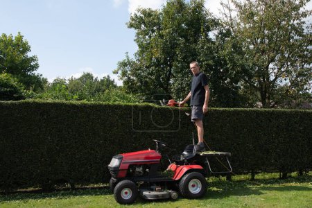 Ein Gärtner oder Arbeiter verwendet Ständer, um Heckenscheren mit Benzin zu schneiden, während er auf einem Traktormäher statt einer Leiter steht, qualitativ hochwertiges Foto. Hochwertiges Foto