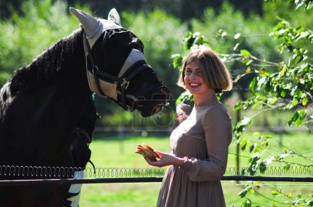 Téléchargez les photos : Femme blonde confiante nourrir un cheval parmi les grands arbres en été, la campagne, rural, scène rustique, photo de haute qualité - en image libre de droit