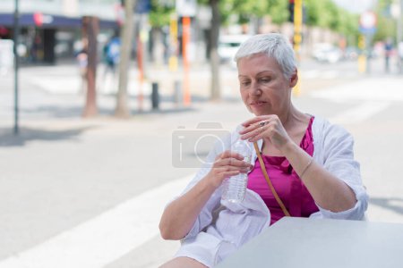 Foto de Una mujer caucásica de sesenta años con bob cortado en ropa de verano elegante beber agua de la botella de plástico en la mesa de la cafetería en la ciudad, Foto de alta calidad - Imagen libre de derechos