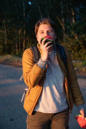 Blonde girl with short hair like a boy,appetizingly bites a large piece of watermelon and laughs, portrait at sunny golden hour at sunset in the forest while walking with friends, high quality photo
