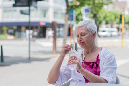 Foto de Una mujer caucásica de sesenta años con bob cortado en ropa de verano elegante beber agua de la botella de plástico en la mesa de la cafetería en la ciudad, Foto de alta calidad - Imagen libre de derechos