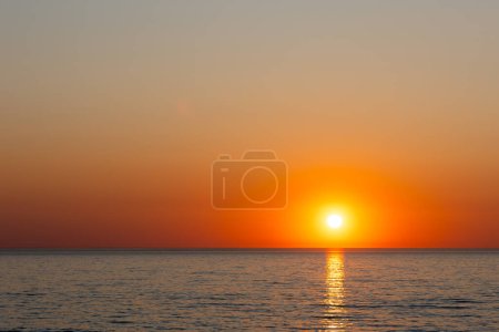 Photo for A large orange sun setting over a calm sea. Static shot taken during a cloudless evening - Royalty Free Image