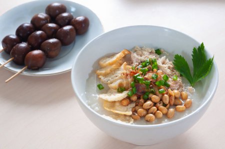 Bubur Ayam Spesial contains white rice porridge with side dishes of fried chicken slices, fried soybeans, emping crackers and sprinkled with fried onions and celery. Served with Cooked quail eggs skewered like satay