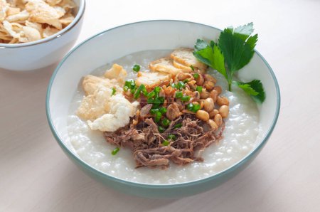 Bubur Daging Krawu Gresik enthält weißen Reisbrei mit Beilagen aus geschreddertem Rindfleisch, gebratenen Sojabohnen, leerem Cracker und bestreut mit gebratenen Zwiebeln und Sellerie