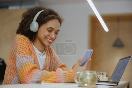 A happy woman, wearing headphones, is seated at a table with laptop and cell phone, smiling as she uses audio equipment and a personal computer
