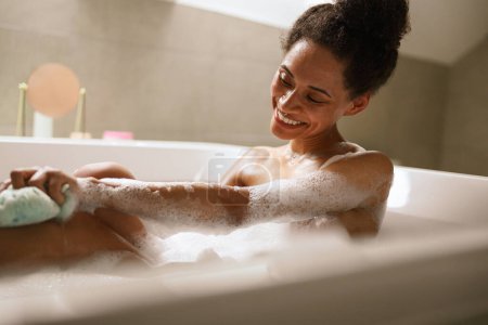 Die Frau badet fröhlich im Whirlpool und wäscht sich mit einem Schwamm Brust und Ellbogen. Sie genießt den Komfort und Spaß der Freizeit, umgeben von weicher Bettwäsche und blättert in einem Magazin