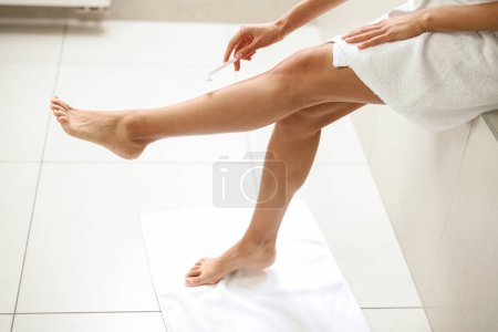A woman, clad in a towel, is delicately shaving her legs with fluid wrist movements, thumb supporting the razor. Her knee rests on the flooring, sleeve pulled up to the elbow