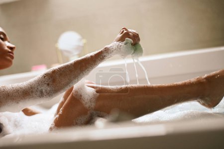 Eine Frau badet fröhlich in einer Badewanne und hält einen Schwamm in der Hand. Ihr Ellbogen ruht am Rand der Wanne, während ihr Bein in Richtung Fenster mit einer Holzpflanze in der Nähe gestikuliert.