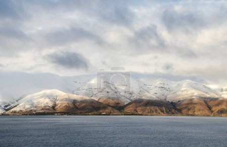 Calm water of Lake Sevan at the foot of the snow-capped mountain peaks under blue sky with low clouds. Amazing natural landscape in Armenia