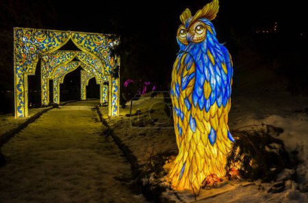 Scène nocturne magique représentant une sculpture majestueuse illuminée de hibou décorée de motifs bleus et jaunes contre un paysage enneigé