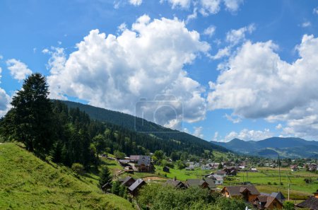 Serene village landscape with houses nestled on the valley among rolling green hills with mountain ridge on background under blue sky with clouds. Verkhovyna, Carpathian Mountains, Ukraine