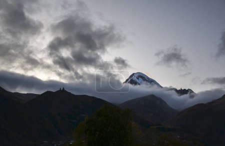 Photo for View from Stepantsminda to silhouette of the Gergeti Monastery against the backdrop of majestic mount Kazbek with snow-capped peak, veiled in misty clouds. Travel to Georgia - Royalty Free Image