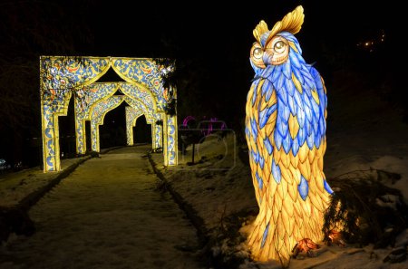 Magical night scene depicting an illuminated majestic owl sculpture decorated with blue and yellow patterns against a snowy landscape