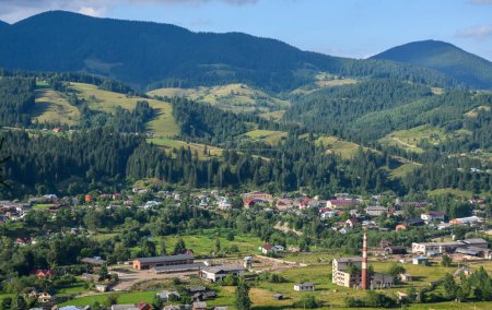 Panoramic view of Verkhovyna village in the lush green valley nestled among rolling forested hills and majestic mountains in the distance. Carpathians, Ukraine 