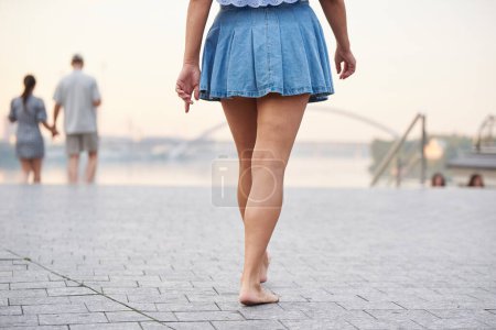 An adult woman in a denim skirt walks barefoot along the city embankment in Kyiv