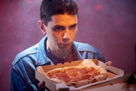 Young calm guy with pepperoni pizza in a cardboard box in the evening in a pizzeria.