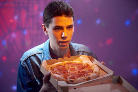 Joven chico tranquilo con pizza de pepperoni en una caja de cartón por la noche en una pizzería.